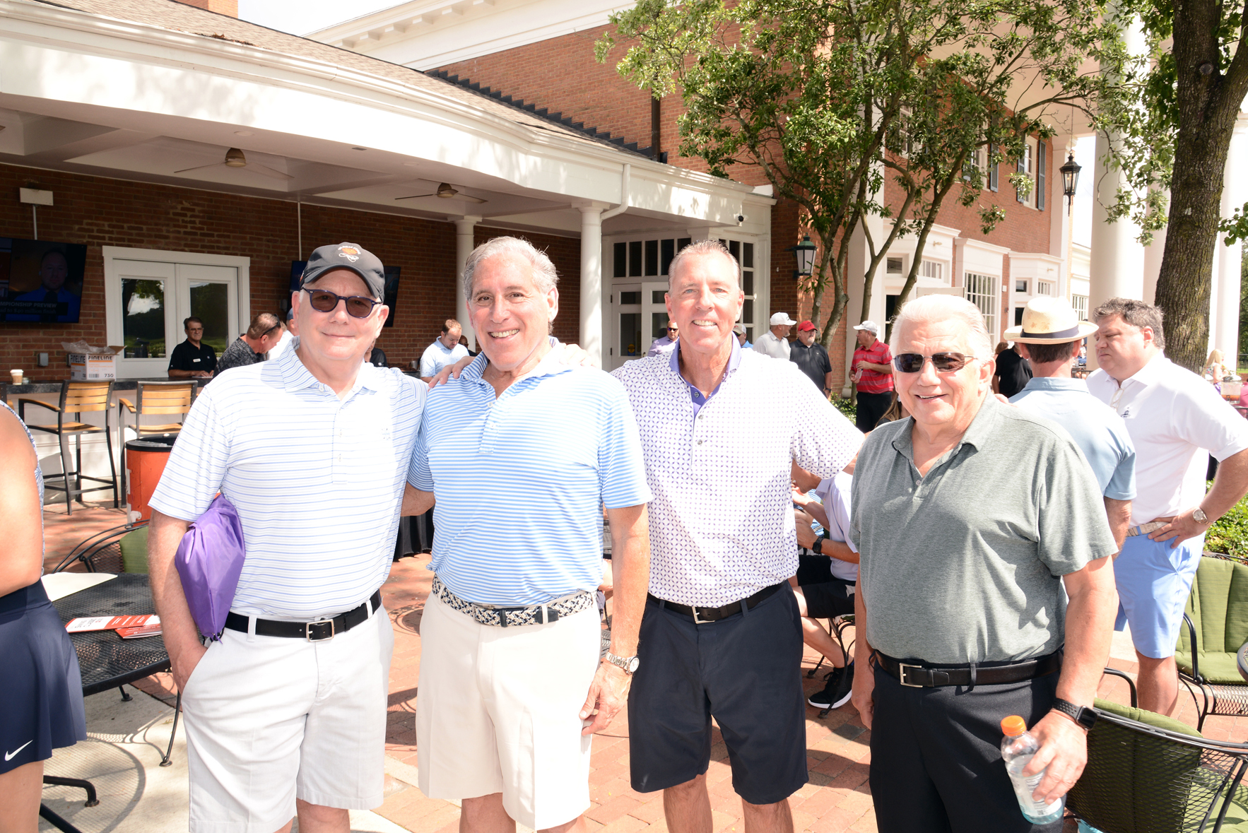 Four people standing outdoors in front of a building with white columns and a brick facade. Faces are obscured. They are dressed casually in polo shirts and shorts. Other attendees are visible in the background, sitting at tables or standing, suggesting a social gathering on a sunny day.