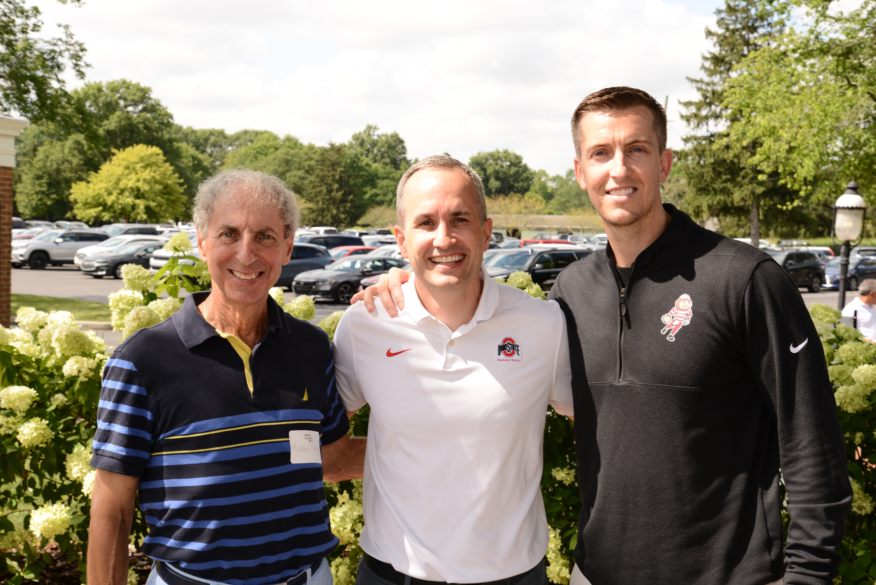 Three individuals standing outdoors with faces blurred, posing in front of greenery and parked cars. One wears a striped polo shirt, another a white Ohio State University polo, and the third a black jacket with an emblem.