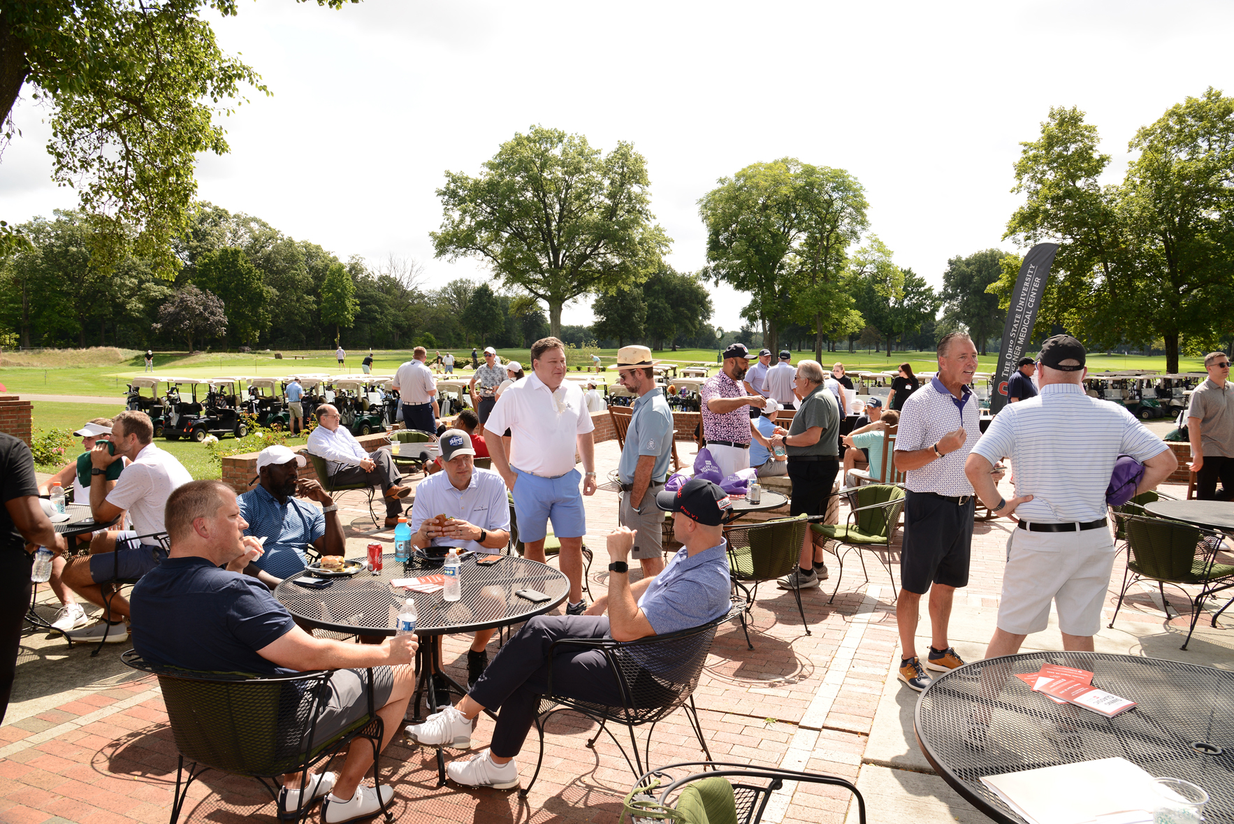 Group of people socializing on a shaded patio at a golf course, with tables, chairs, food, drinks, and golf carts visible in the background.