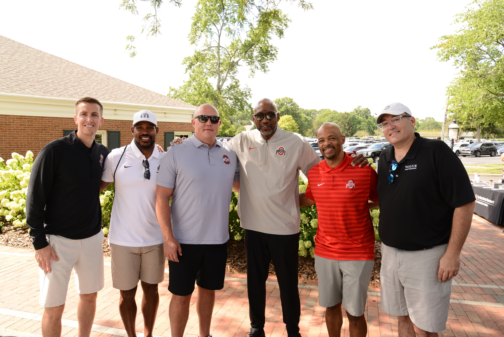 Six individuals standing outdoors on a brick pathway, dressed in casual clothing including shorts and polo shirts with logos. Greenery and a building are visible in the background.