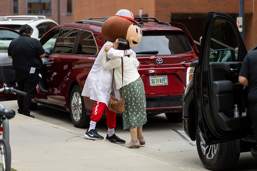 Brutus Buckeye in a lab coat hugging an individual outside a building.
