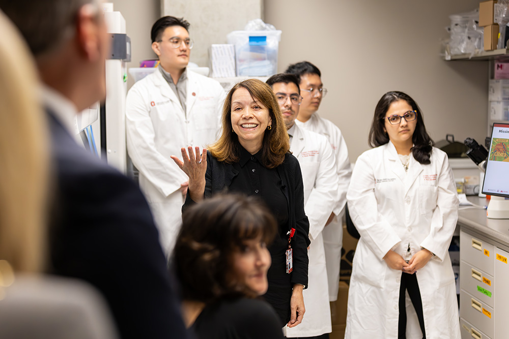 Women in black talking with others while four students in lab coats stand behind her