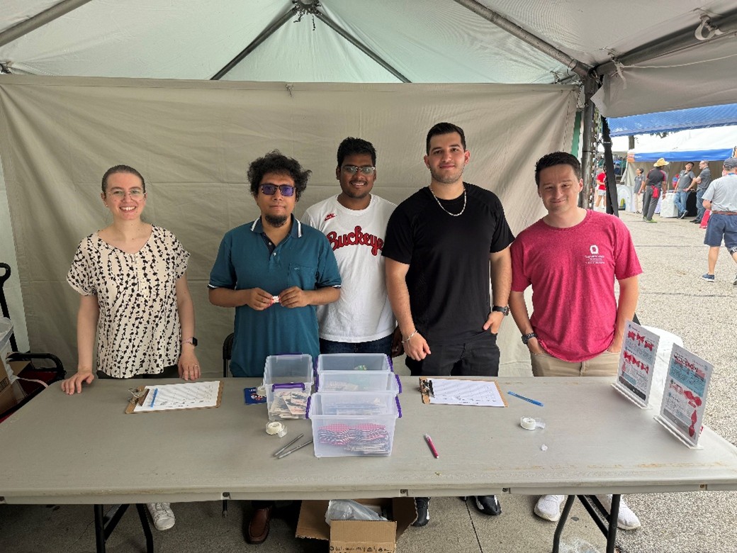 Five people stand behind a table under a tent at an outdoor event, with clipboards, pens, and clear supply bins arranged on the table.