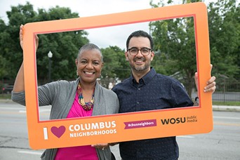 Two people standing outdoors holding a large orange photo frame that reads ‘I ❤️ Columbus Neighborhoods’ and ‘WOSU Public Media.’