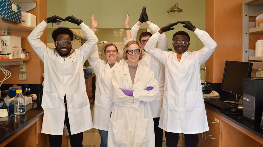 Five people in lab coats standing in a laboratory, posing with their arms raised to form shapes.