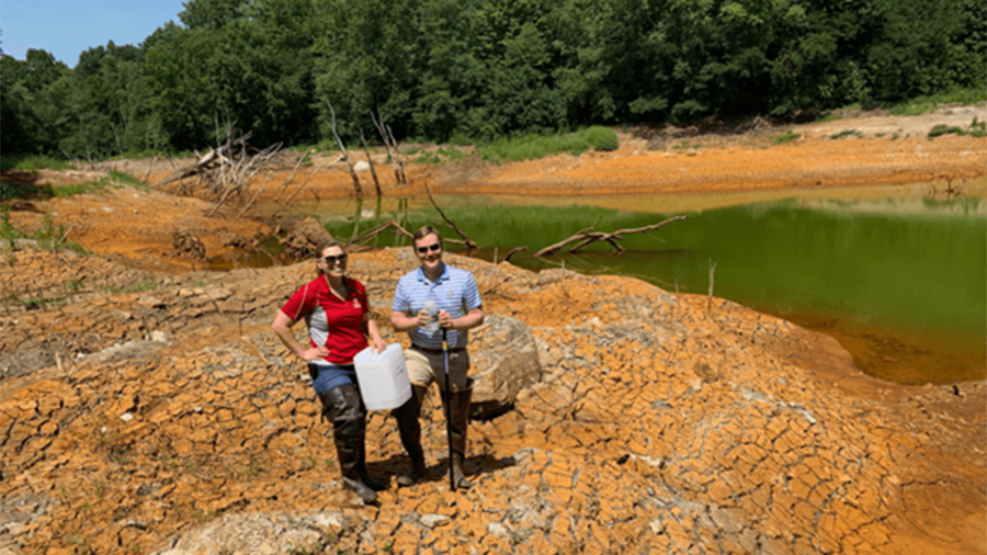 Two people standing on cracked, dry ground near a small green pond in a drought-affected area, holding water sampling equipment.