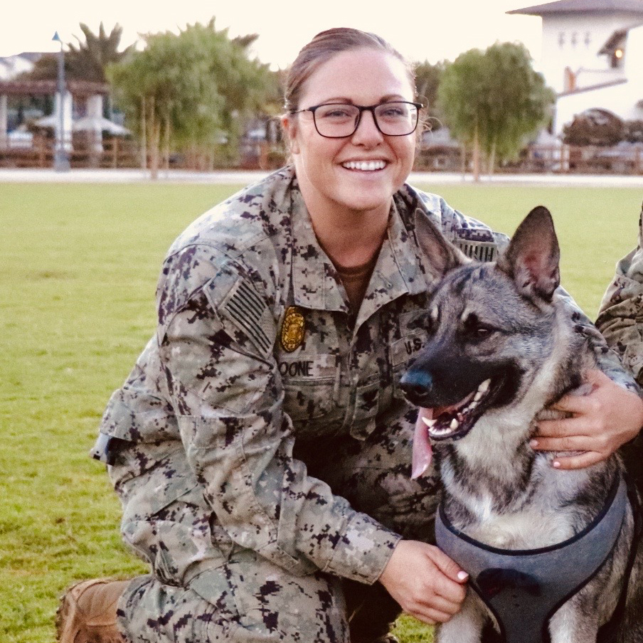 Elizabeth in a camouflage military uniform kneels on grass, holding a working dog wearing a harness, with buildings and trees in the background.