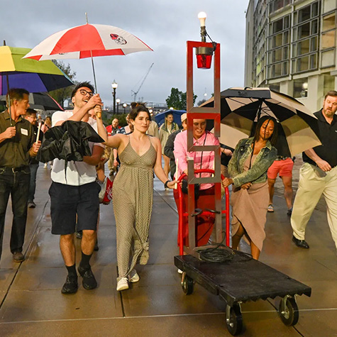 A group of Ohio State theatre students and teachers walk outside carrying umbrellas and pushing a cart with a light