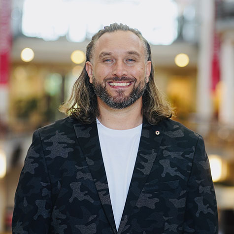 A man with long, brown hair stands and smiles wearing a black jacket and a white t-shirt under it