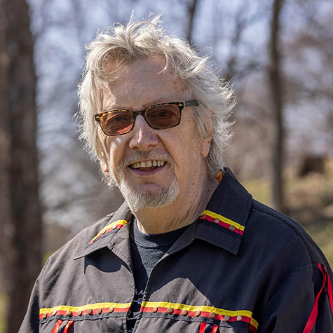 A man with white hair and wearing sunglasses stands outside on a sunny day with tall trees behind him