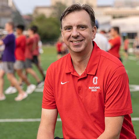 Photo of Ohio State Marching Band Director as he sits outside wearing a red polo shirt while the band practices behind him 