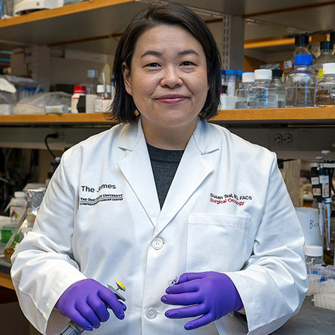 Photo of a woman wearing a white lab jacket and purple gloves as she stands in a research laboratory in front of shelves with bottles