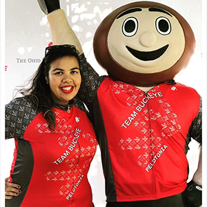 Selena Browning, a woman with dark hair, wearing a Pelotonia Team Buckeye shirt smiles and stands next to a statue of Brutus Buckeye, who is also wearing a Team Buckeye shirt