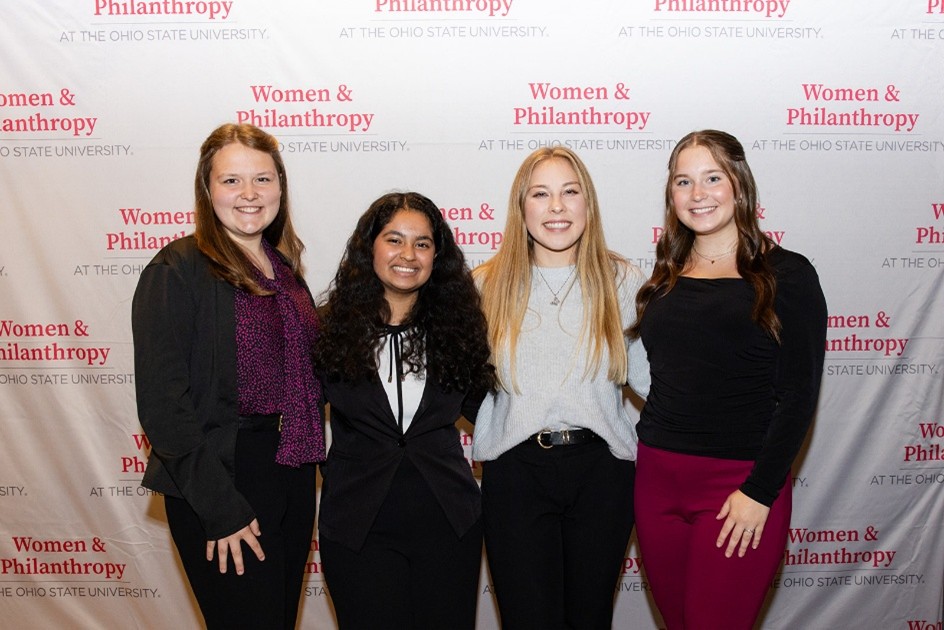 Four students standing in front of a Women & Philanthropy backdrop posing for a photo.