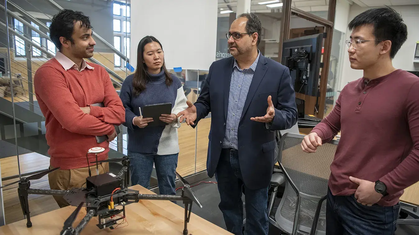 engineering students talk with professor in front of a table with a drone and parts sitting on top