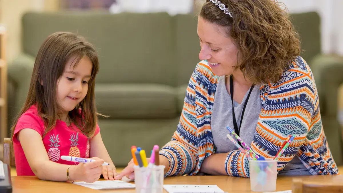 woman in bright colored sweater working with a young girl on an art project at a table