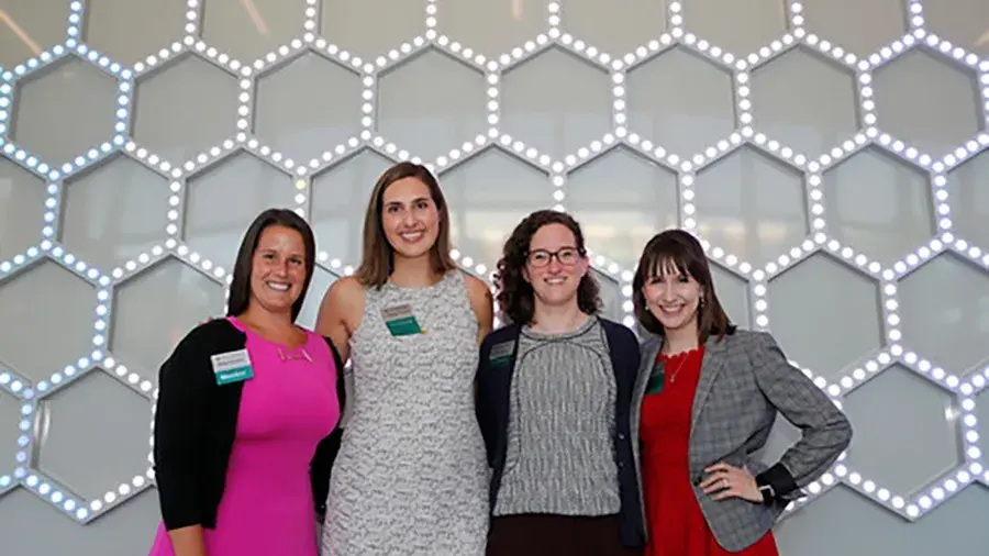 four women in dresses and jackets posing for a picture against a honeycomb textured wall