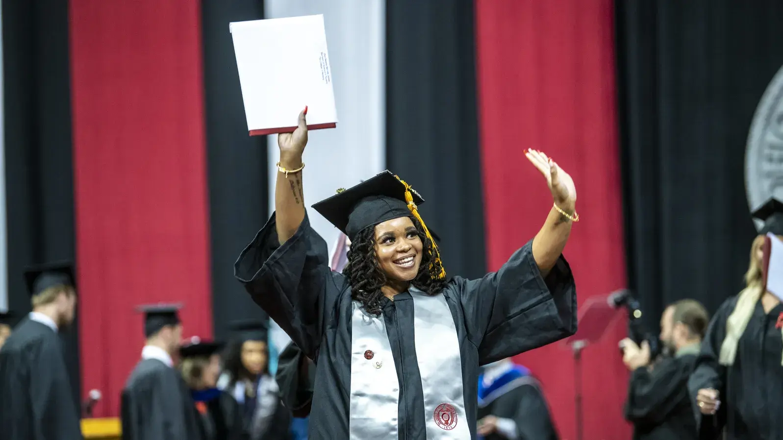 woman in graduation cap and gown waves to the audience after receiving her diploma