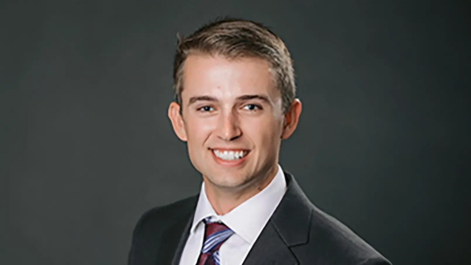 Adam Kaye wearing a suit posting in front of a gray backdrop