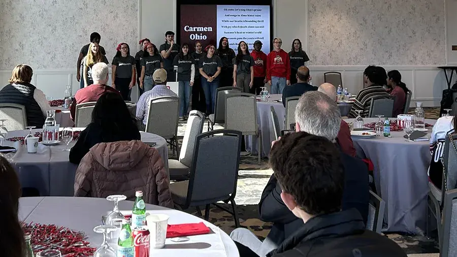 A group of Ohio State students stand and sing Carmen Ohio in front of a room full of people who are seated at tables