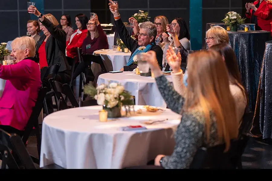 A group of women sitting at tables raise their glasses in a celebration toast