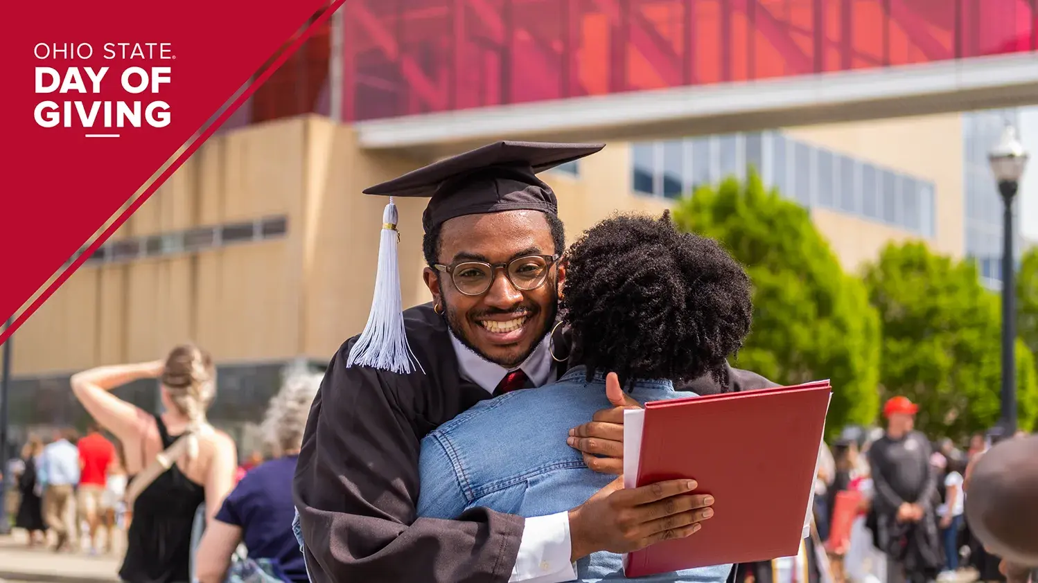 A graduate wearing a cap and gown smiles and hugs a person while holding a diploma