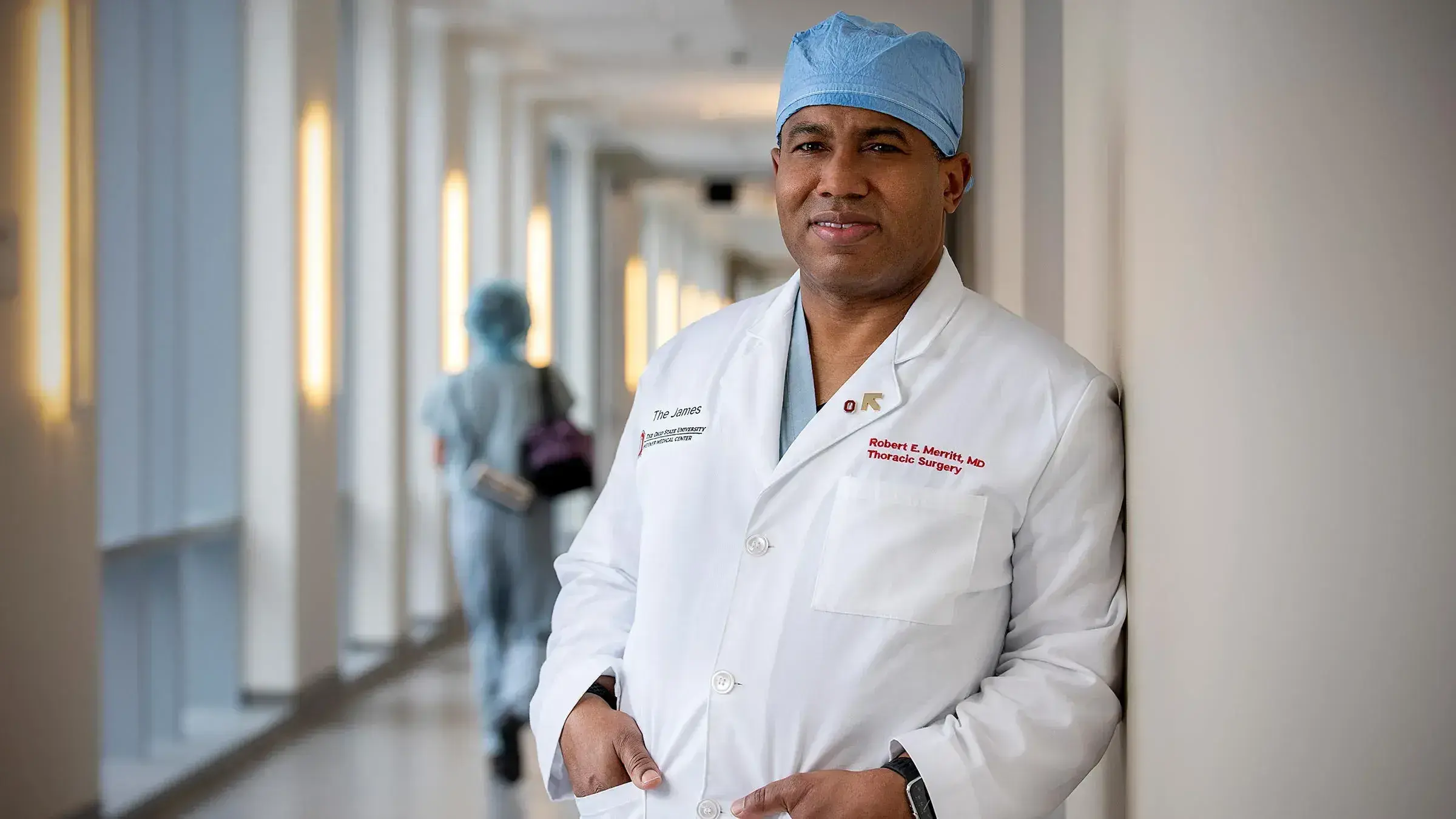 A doctor wearing a scrub hat and white lab jacket stands and smiles