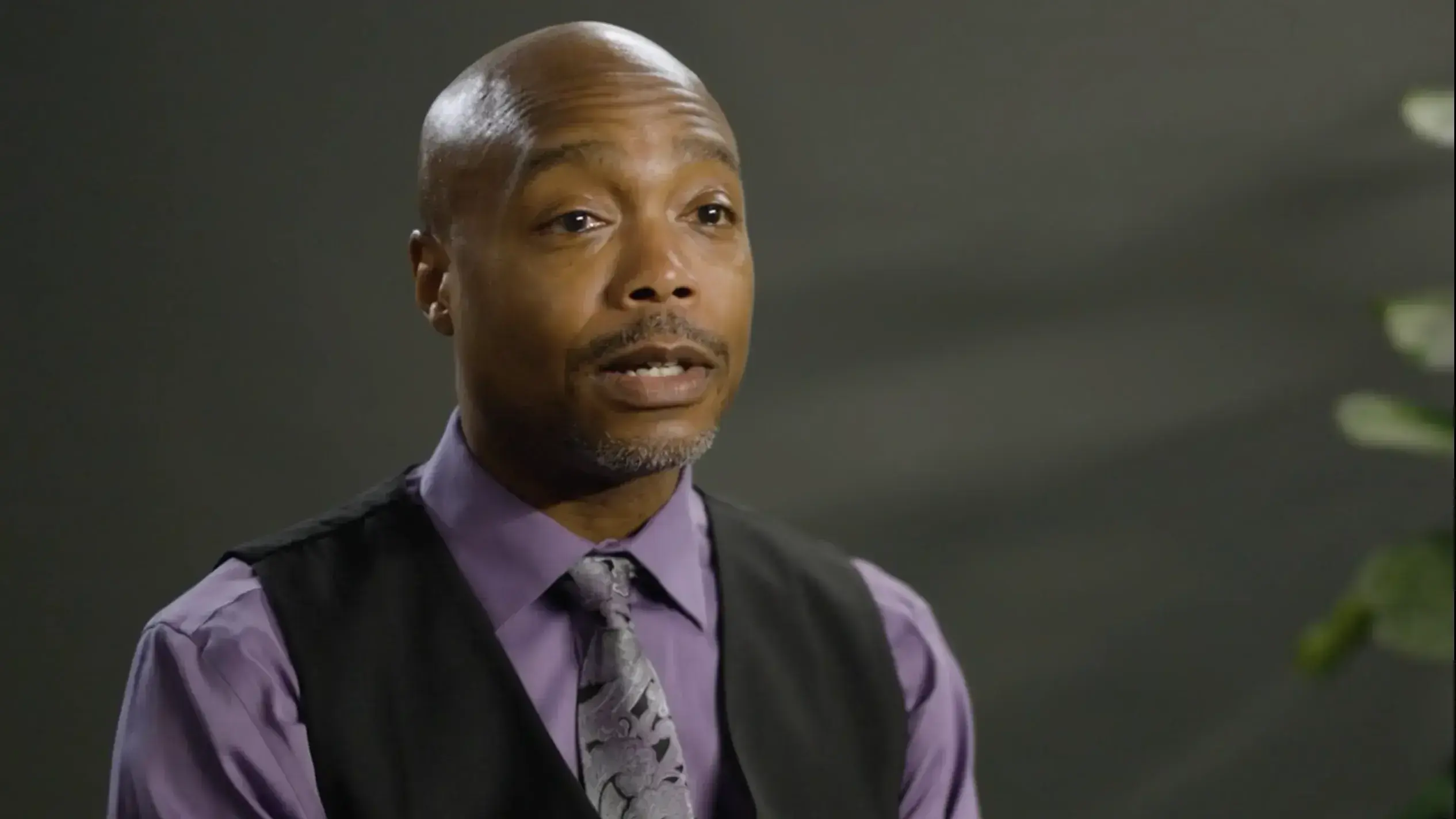 A man wearing a purple shirt and black vest appears to be talking