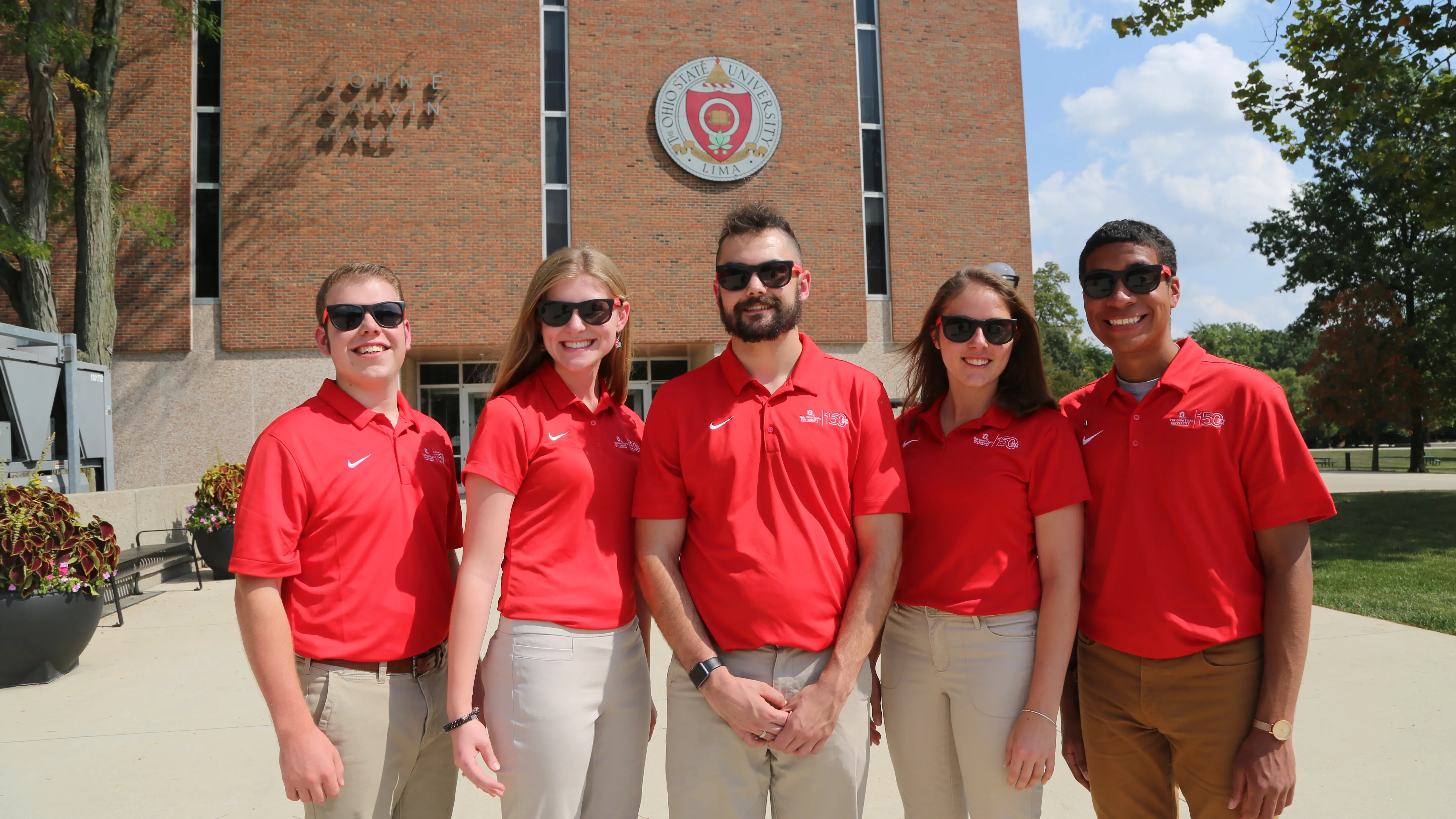 A group of Ohio State students wearing red shirts and sunglasses stand outside on a bright, sunny day and smile