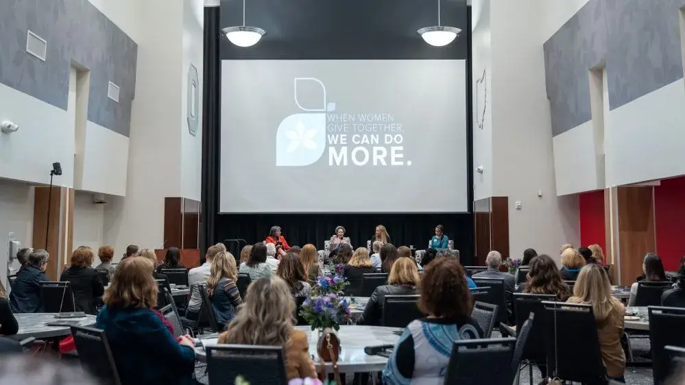 A large group of people sit in a room at round tables listening to a panel of speakers talk.