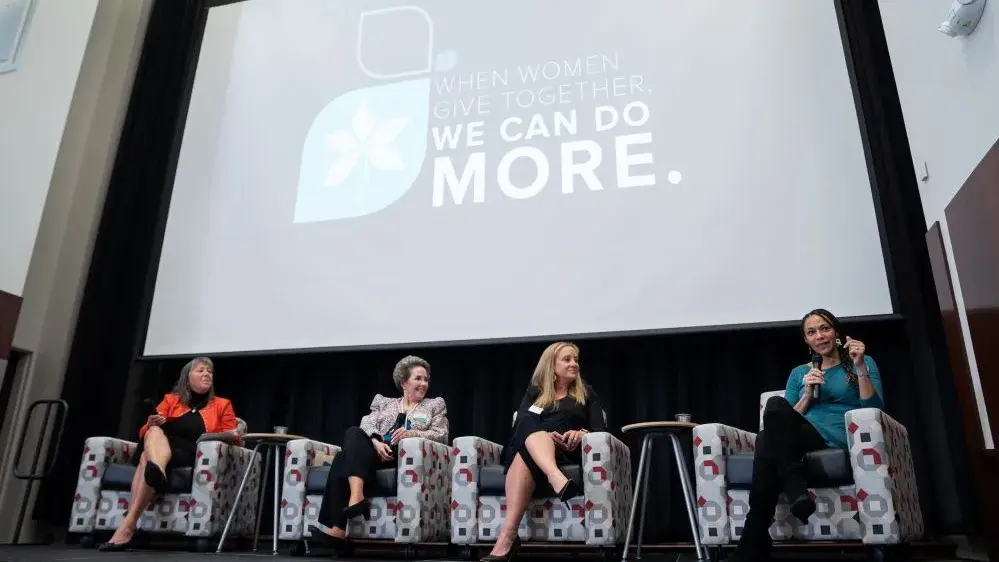 Four women sit in chairs on a stage as part of a speaker panel. Three of them are listening to the other talk.