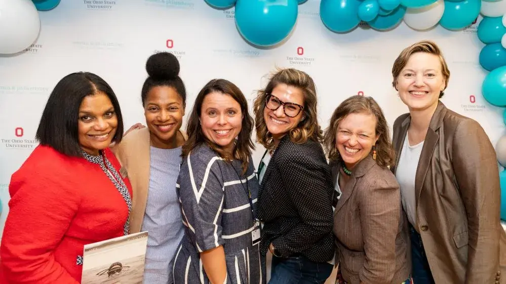 A group of six women stand together with big smiles and some are laughing