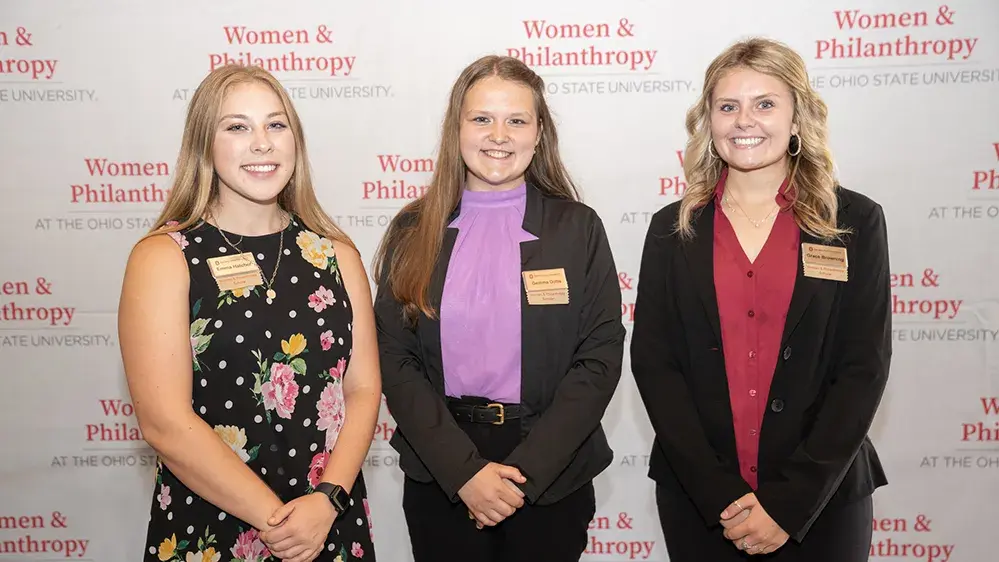 Three young women dressed in business casual clothing stand and smile