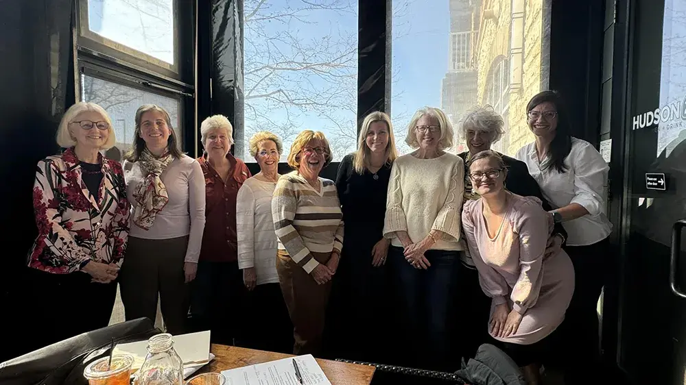 A group of women stand together and smile in front of large windows and a table in front of them