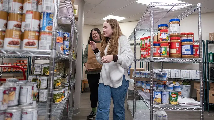 Two students in the Monda Resource Center surrounded by shelves with food donations.
