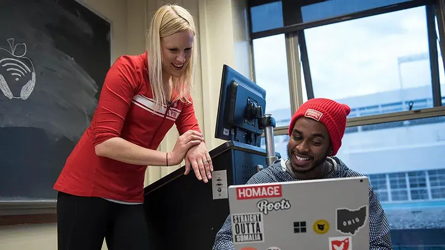 A person in a red top leans over a desk helping another person who is seated at a computer with a laptop covered in stickers. A chalkboard and a window are visible in the classroom setting.