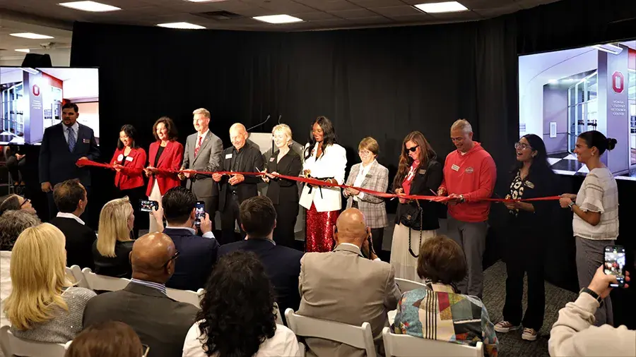A group of people stand in a row on a stage holding a long red ribbon during a ribbon‑cutting ceremony. An audience sits facing the stage, and large screens on either side display images of a building interior with an Ohio State “O” symbol.