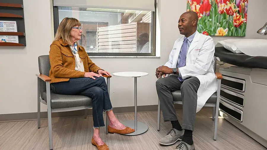 A patient sits across from a medical professional in an exam room, having a conversation beside a small round table.