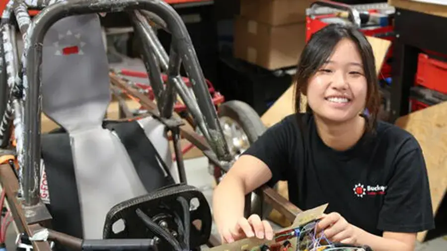 Student working on wiring components next to a small open‑frame race car in a workshop.