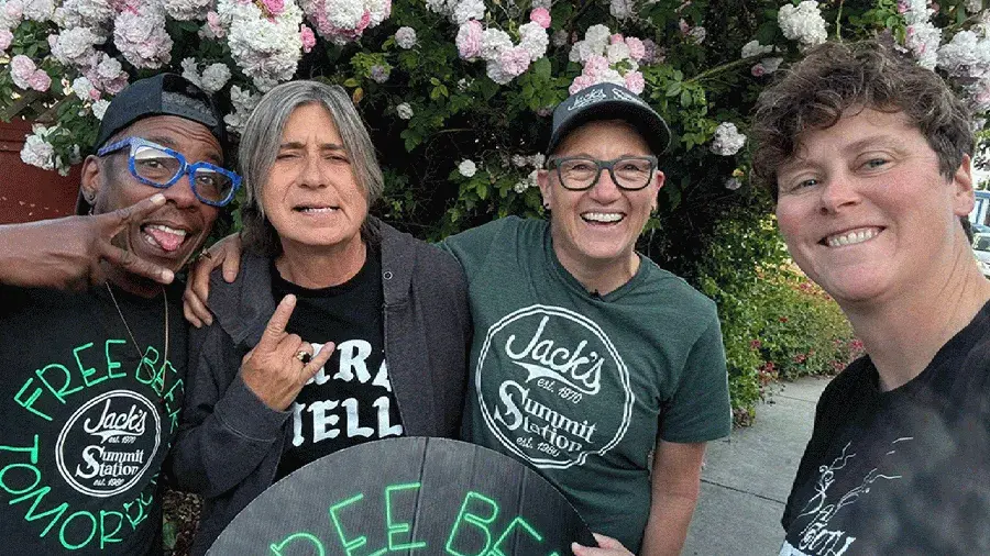 Four people stand close together in front of a flowering shrub, wearing shirts and holding a round sign with bright lettering that reads “FREE BEER TOMORROW.” Some shirts display the text “Jack’s Summit Station.”