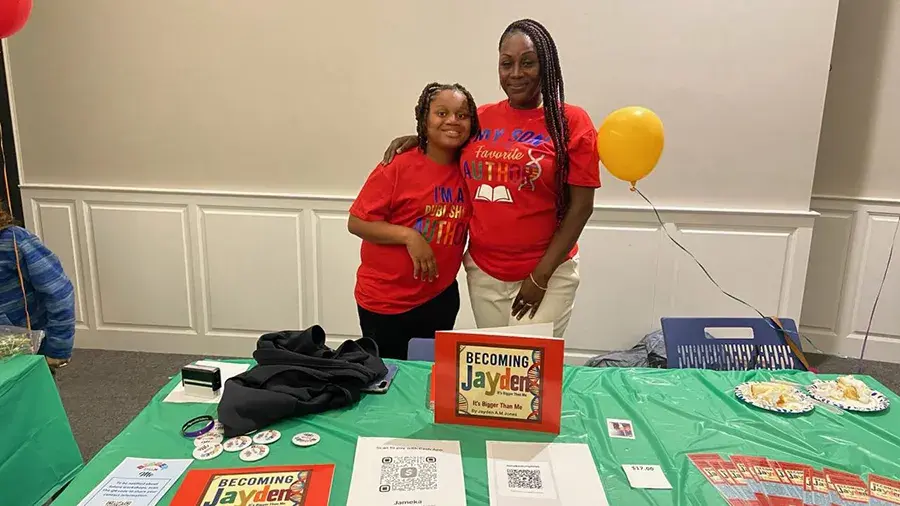 A table with “Becoming Jayden” books, buttons, flyers, and QR codes is set up at an event. Two people in matching red T‑shirts stand behind the table. A yellow balloon and plates of snacks sit to the side.