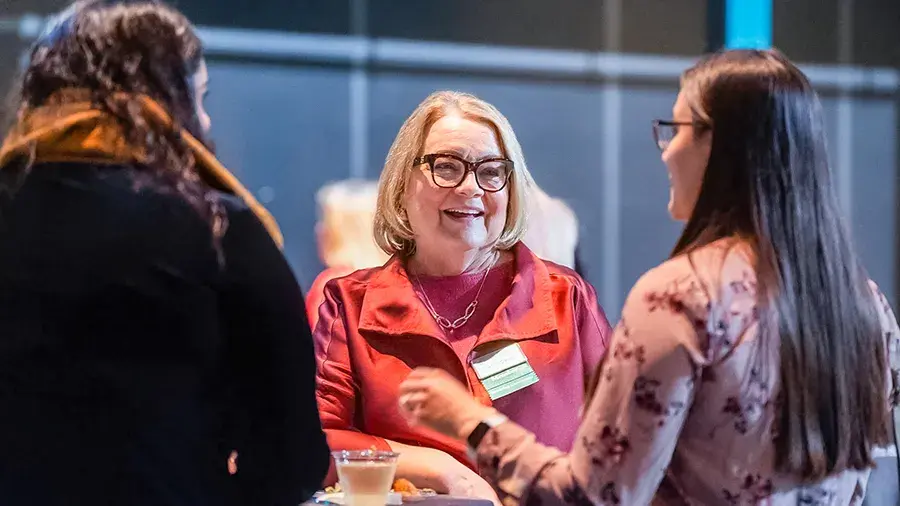 Three people standing and talking together at a social event, gathered around a small table with drinks.