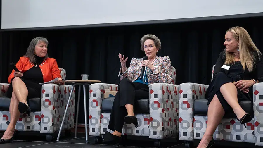 Three speakers seated in patterned armchairs on a stage, engaged in a panel discussion. The person in the center is speaking into a microphone while the others listen.