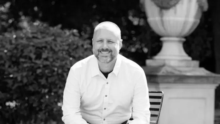A person wearing a long‑sleeve button‑down shirt sits on an outdoor bench in front of a stone structure with greenery in the background.