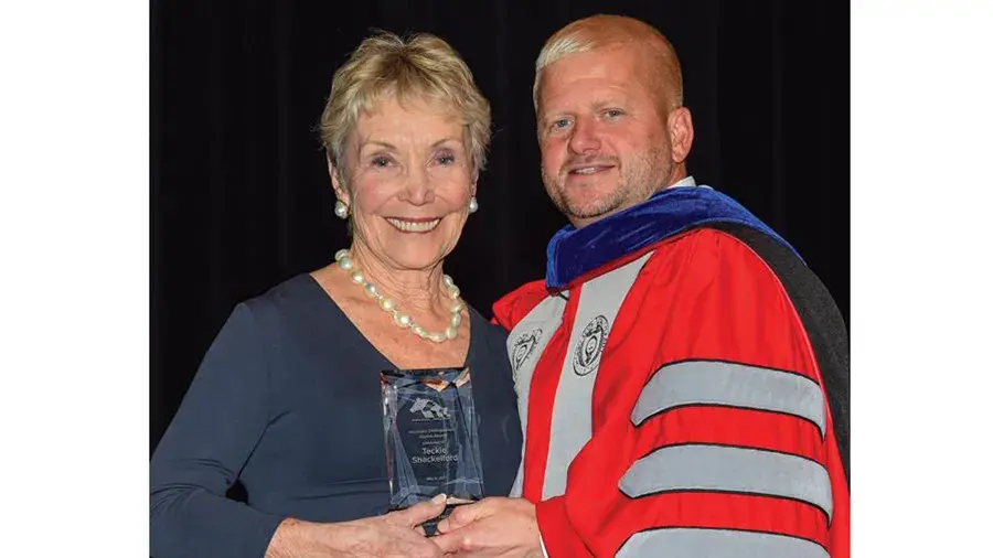 Two people standing together on a stage, holding an award plaque between them; one is dressed in formal attire and the other in an academic robe.