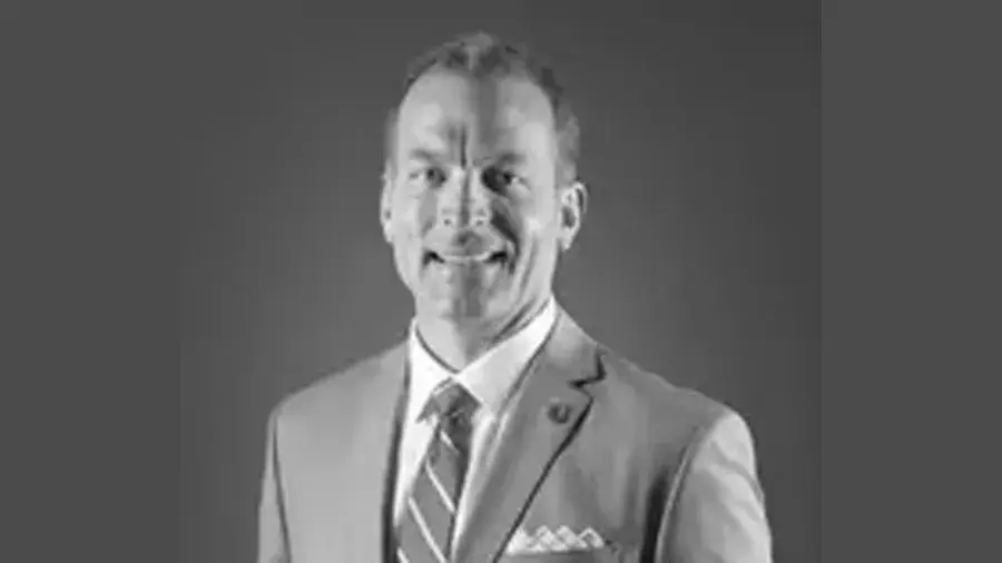 A person wearing a suit jacket, dress shirt, and striped tie is shown in a formal portrait against a plain background. A pocket square is visible in the jacket pocket.