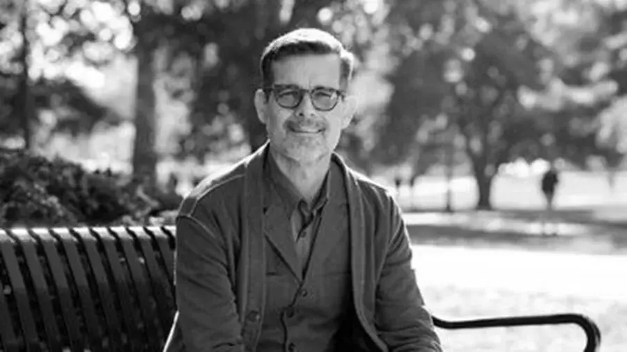 A person sits on an outdoor park bench wearing layered clothing, with trees and open grassy areas visible in the background.