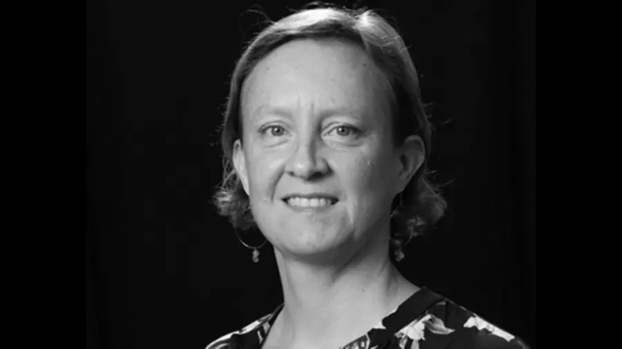 A person with short, wavy hair is shown in a portrait-style image wearing a patterned top against a dark background.