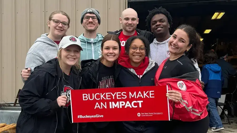 A group of people standing together outdoors holding a red sign that reads ‘Buckeyes Make an Impact’ with the Ohio State University logo and the hashtag #BuckeyesGive.