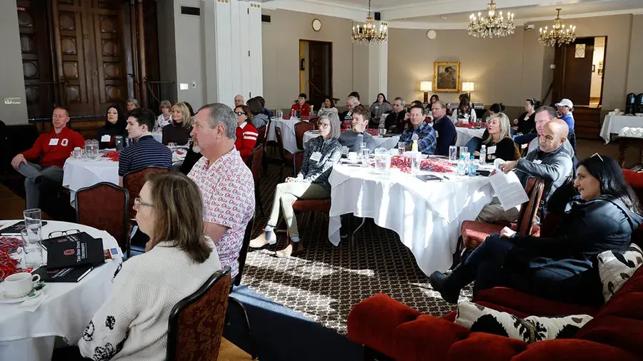A group of people seated at round tables in a large event room, listening to a presentation. The room has chandeliers, white tablecloths, and various materials and drinks on the tables.